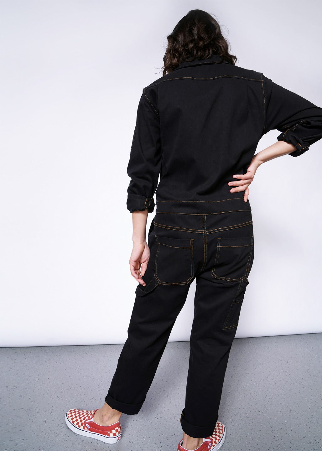Back view of model wearing size small black coverall with orange pop stitch detailing, showing two butt pockets, one side pocket, and hammer loop