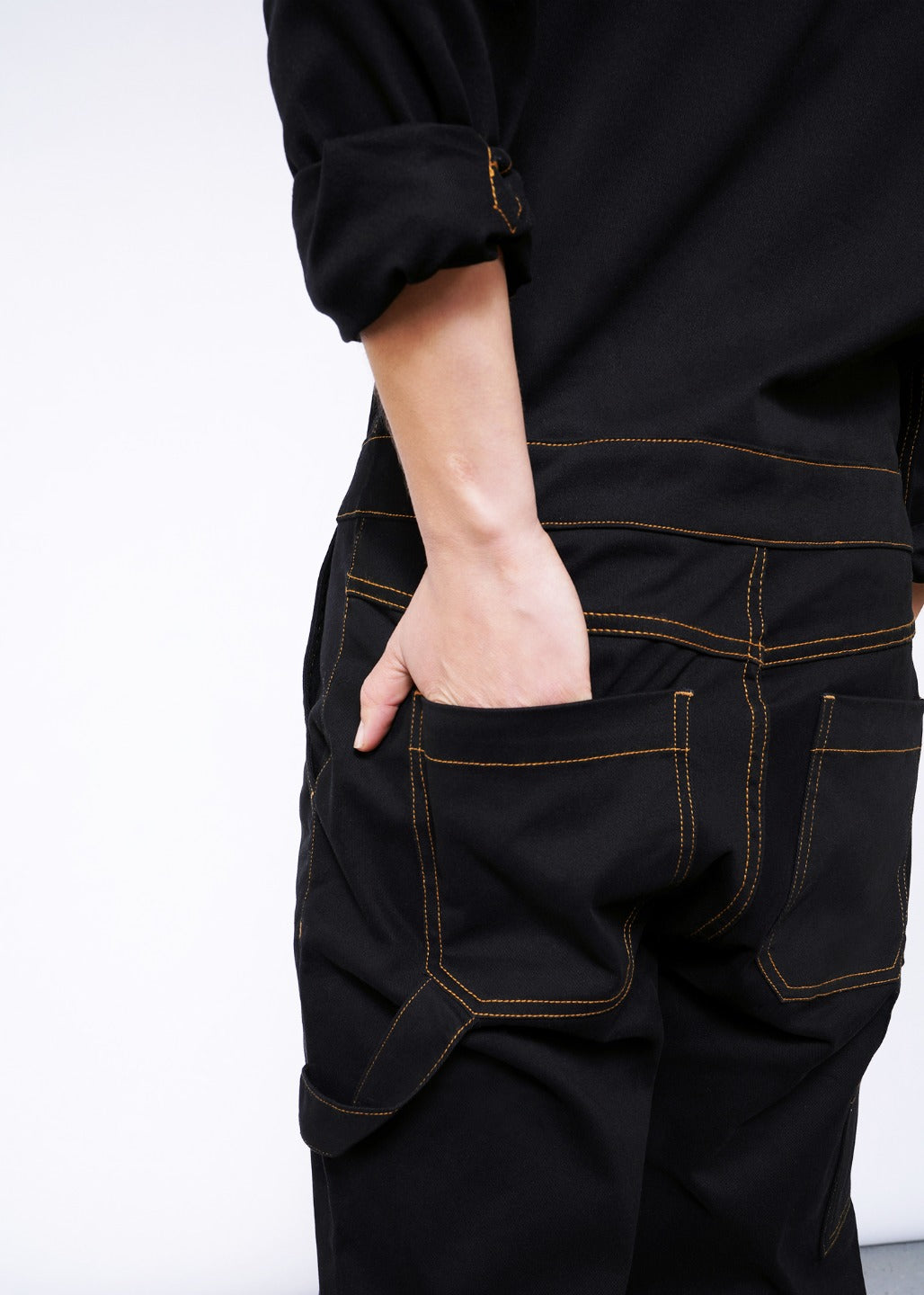 Hand in back pocket of black coverall jumpsuit with orange popstitch detailing