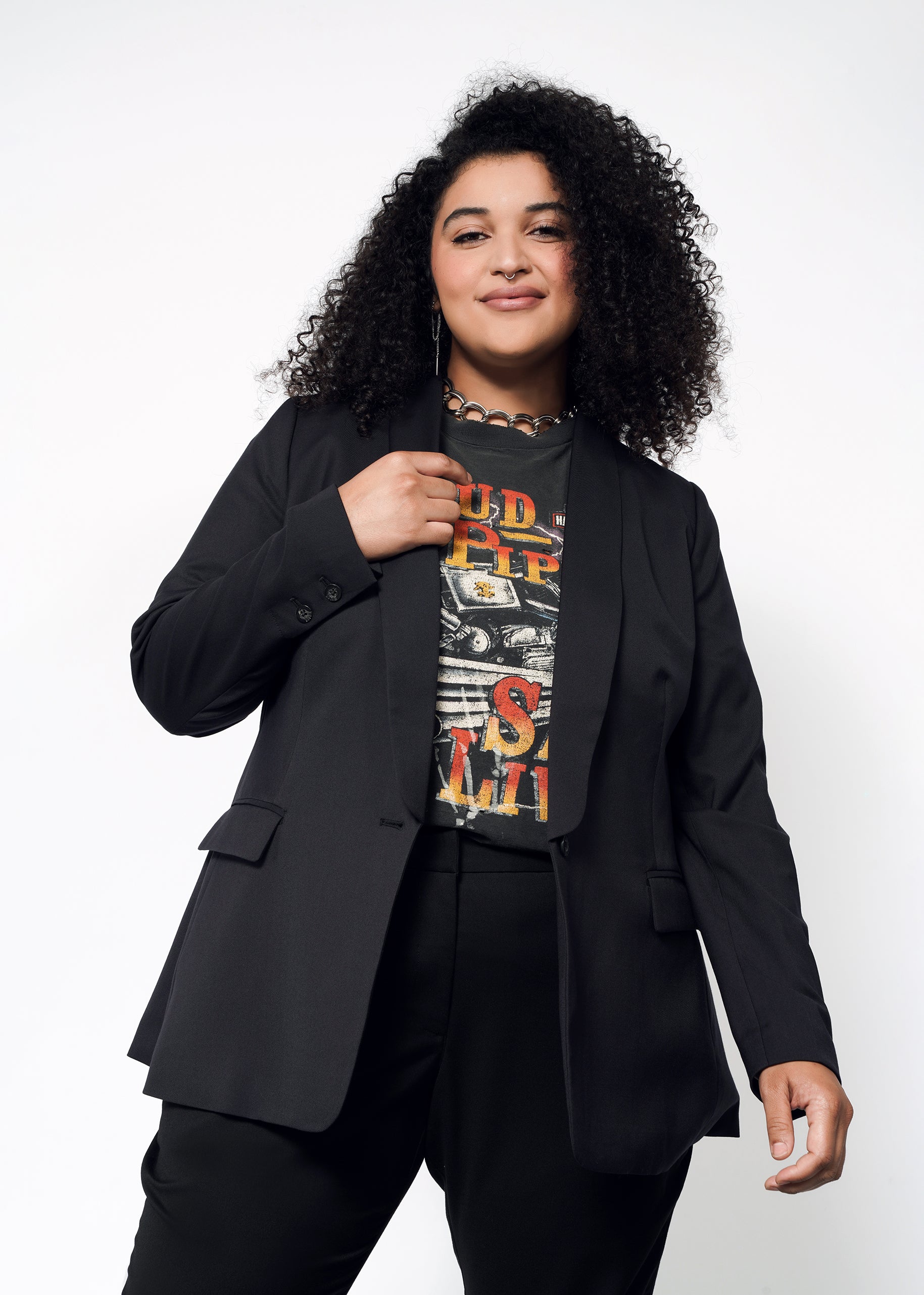 A person with curly hair stands confidently in The Empower Tux Blazer over a graphic T-shirt and black pants, against a plain background.