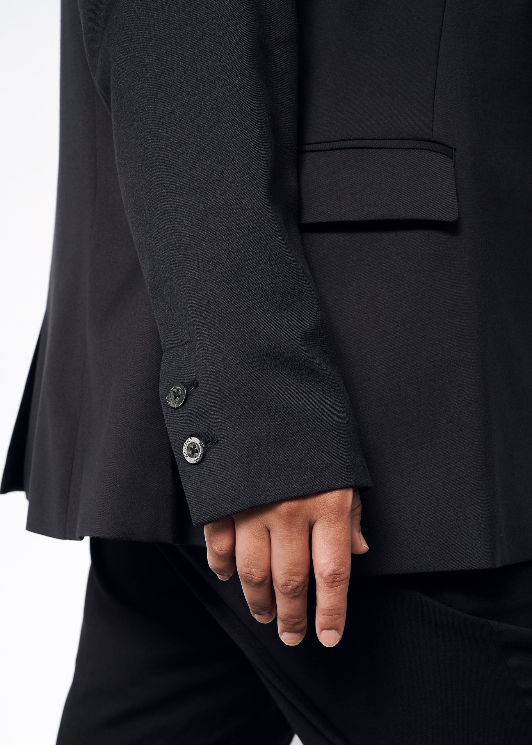 A close-up shows a person wearing The Empower Tux Blazer, featuring a tailored fit with two visible buttons on the sleeve, against a plain and light background. Their hand rests at their side.