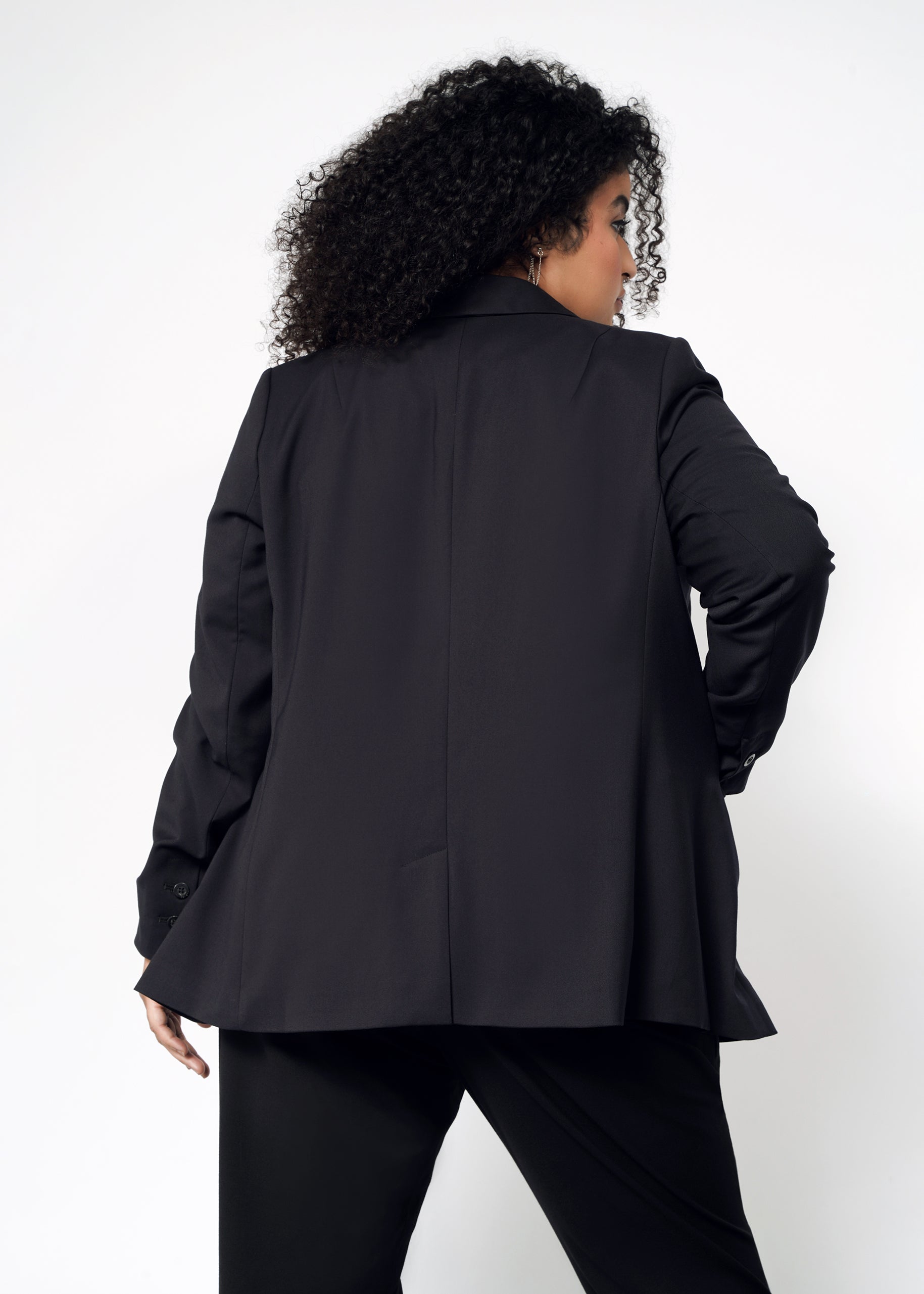 A person with curly hair is seen from behind in a classic pose, wearing The Empower Tux Blazer and matching black pants against a white background. One hand rests confidently on their hip, showcasing this tailored fit ensemble.