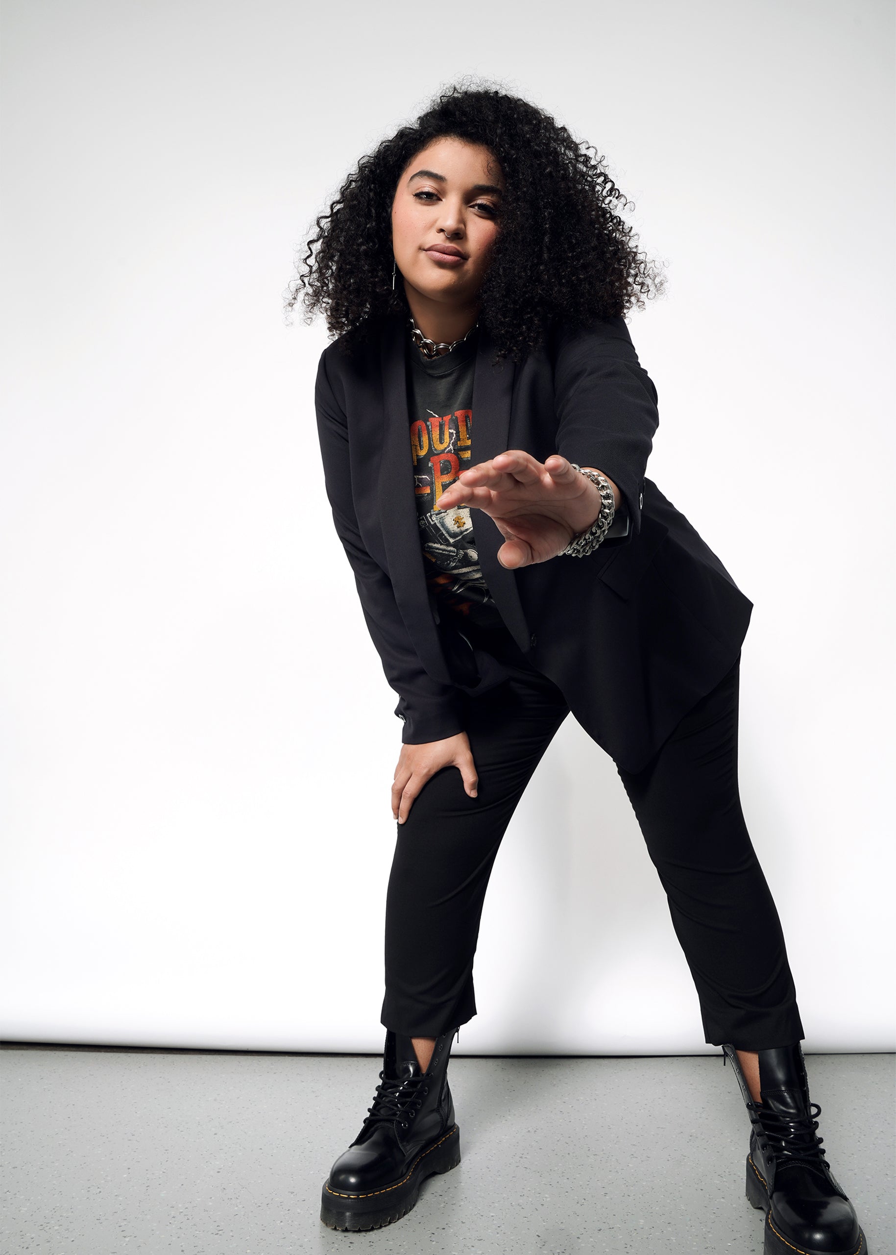 Wearing The Empower Tux Blazer, a person with curly hair in a tailored-fit black outfit and boots confidently poses against a white backdrop, slightly bent forward with one hand extended, exuding a casual and stylish vibe.