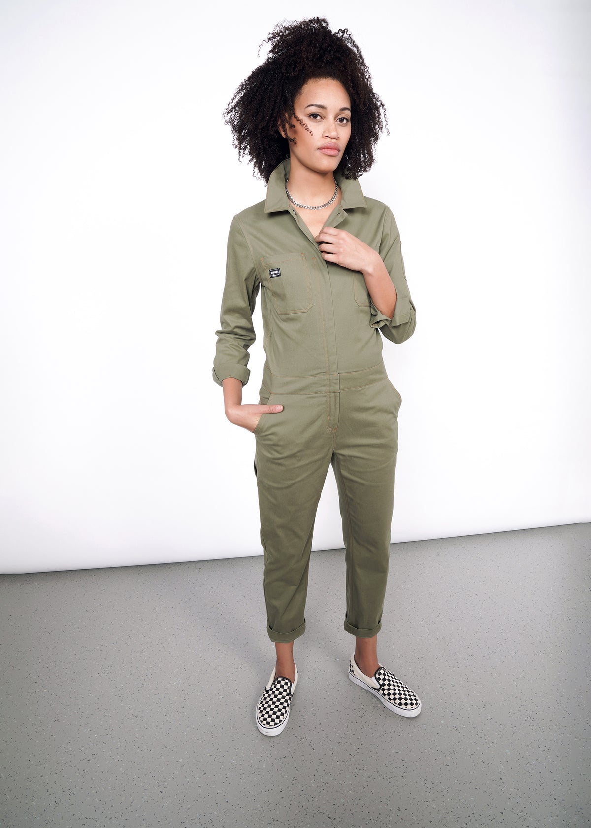 Model wearing olive coveralls slightly unzipped with no shirt underneath, styled with a silver necklace and vans. Coverall legs are cuffed once.