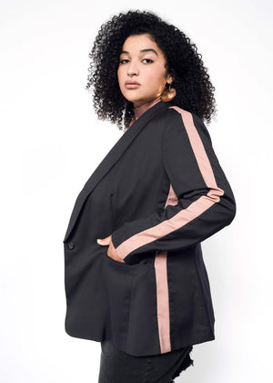 1X=A woman with curly hair models The Empower Colorblock Tux Blazer, featuring its tailored fit and pink tuxedo sleeve stripes. She accessorizes with large hoop earrings, casually placing her hands in the pockets, exuding confidence against a plain white background.