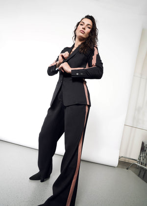 S=A woman confidently poses, adjusting the buttons on her Empower Colorblock Tux Blazer with pink tuxedo stripes. The simple background highlights the blazers tailored fit.