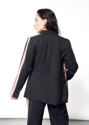 S=A person with dark hair stands against a white backdrop, wearing a stylish black suit featuring pink tuxedo stripes on the sleeves and pants. With a tailored fit, the ensemble includes The Empower Colorblock Tux Blazer, exuding sophistication and modern elegance.