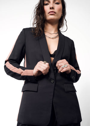 S=A woman poses confidently in The Empower Colorblock Tux Blazer, featuring a black and pink design with a deep neckline and tuxedo stripe. She wears a necklace and rings, hair loose, while gazing down at the camera against a plain white background.