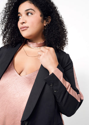 1X=A woman with curly hair looks to the side, wearing The Empower Colorblock Tux Blazer in a tailored fit with pink striped sleeves, paired with a pink satin top, choker, and layered gold necklaces against a plain white background.