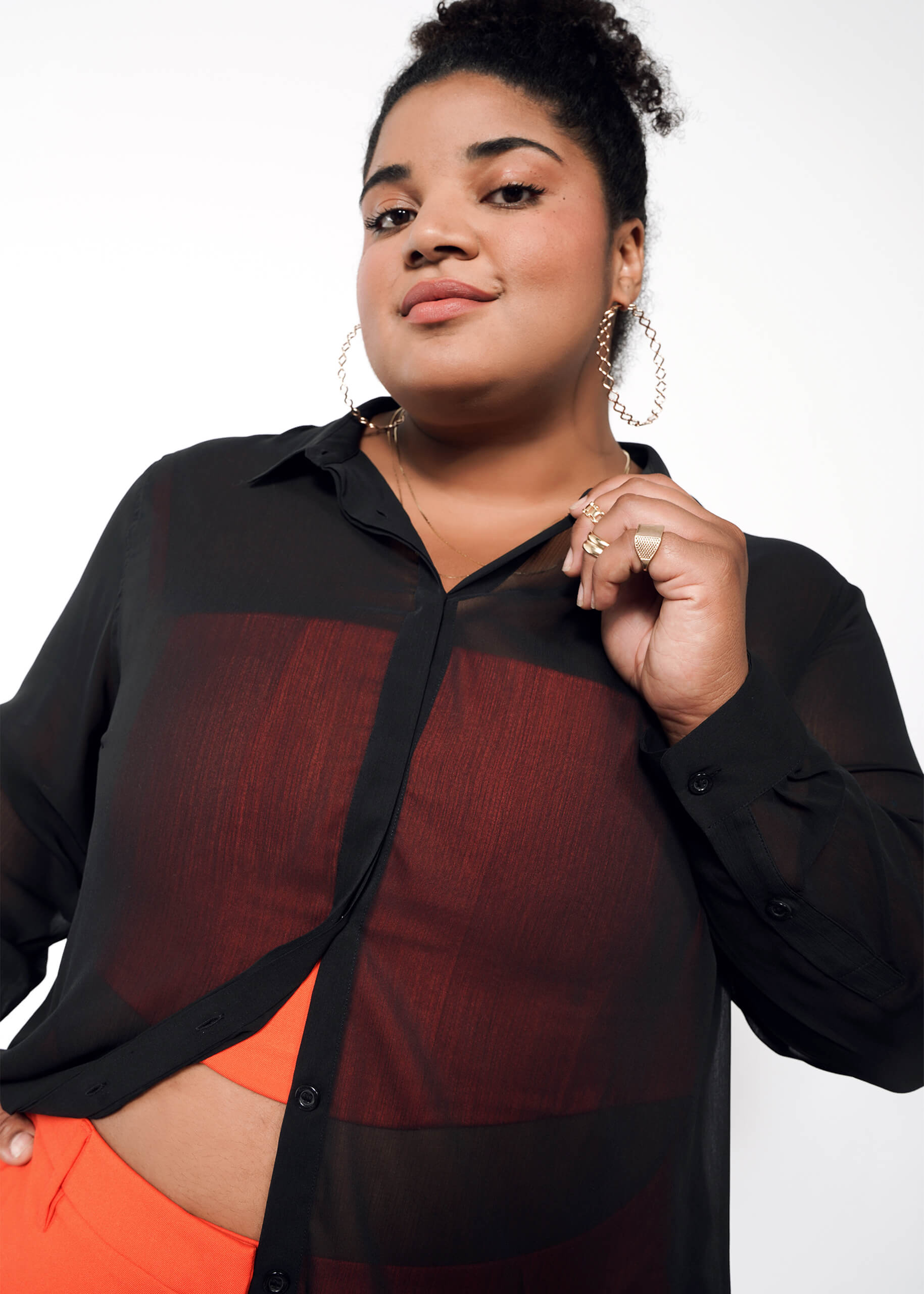 A person with curly hair in a bun wears large hoop earrings and an oversized fit: The Empower Sheer Long Sleeve Button Up over a red shirt, paired with a red skirt. They strike a confident pose, gently pulling on the collar with gold-ringed fingers against a neutral background.