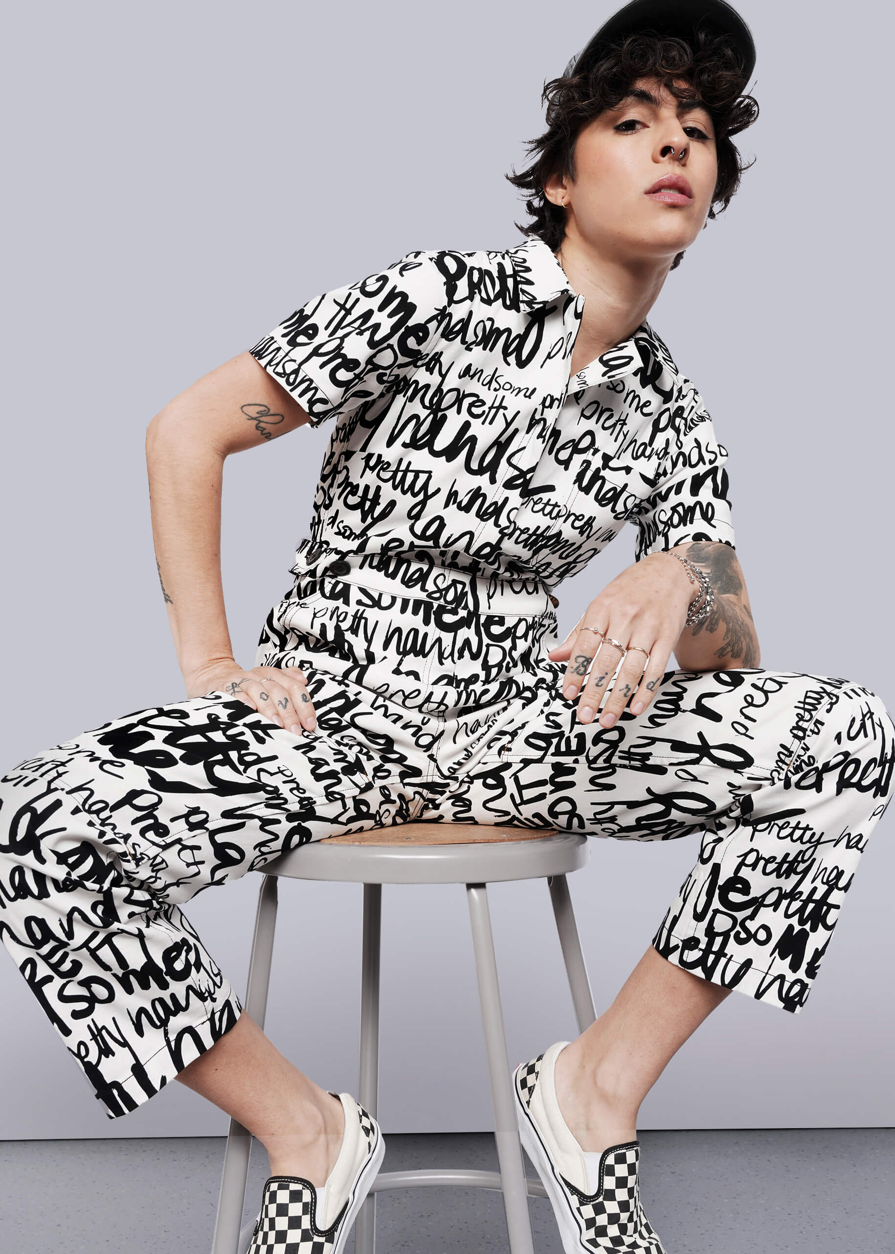 Wearing The Essential High Waisted Coverall in white with bold black handwritten words, a person sits confidently on a stool, styled with a black cap, checkered slip-ons, short curly hair, tattoos, and a self-assured expression.