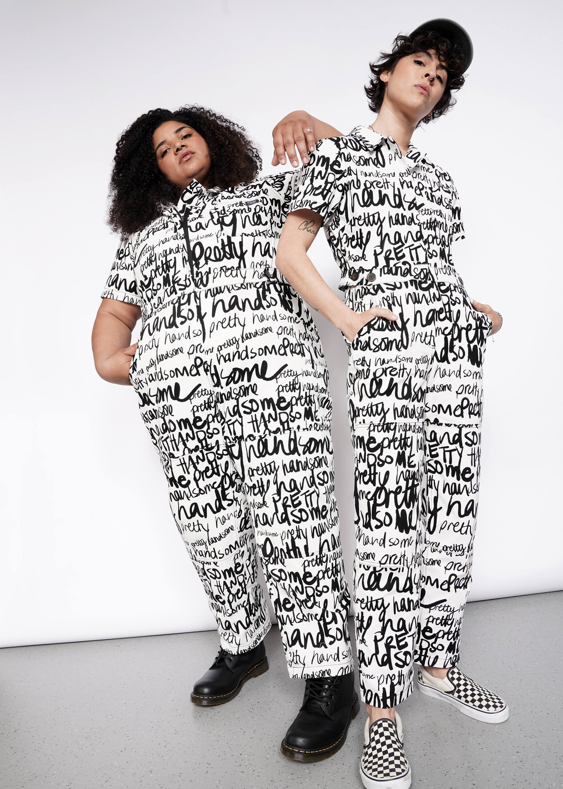 Two people pose against a plain white background in The Essential High Waisted Coverall, featuring a matching white design with bold black handwritten text. One pairs it with boots, the other with checkered slip-ons and a cap.