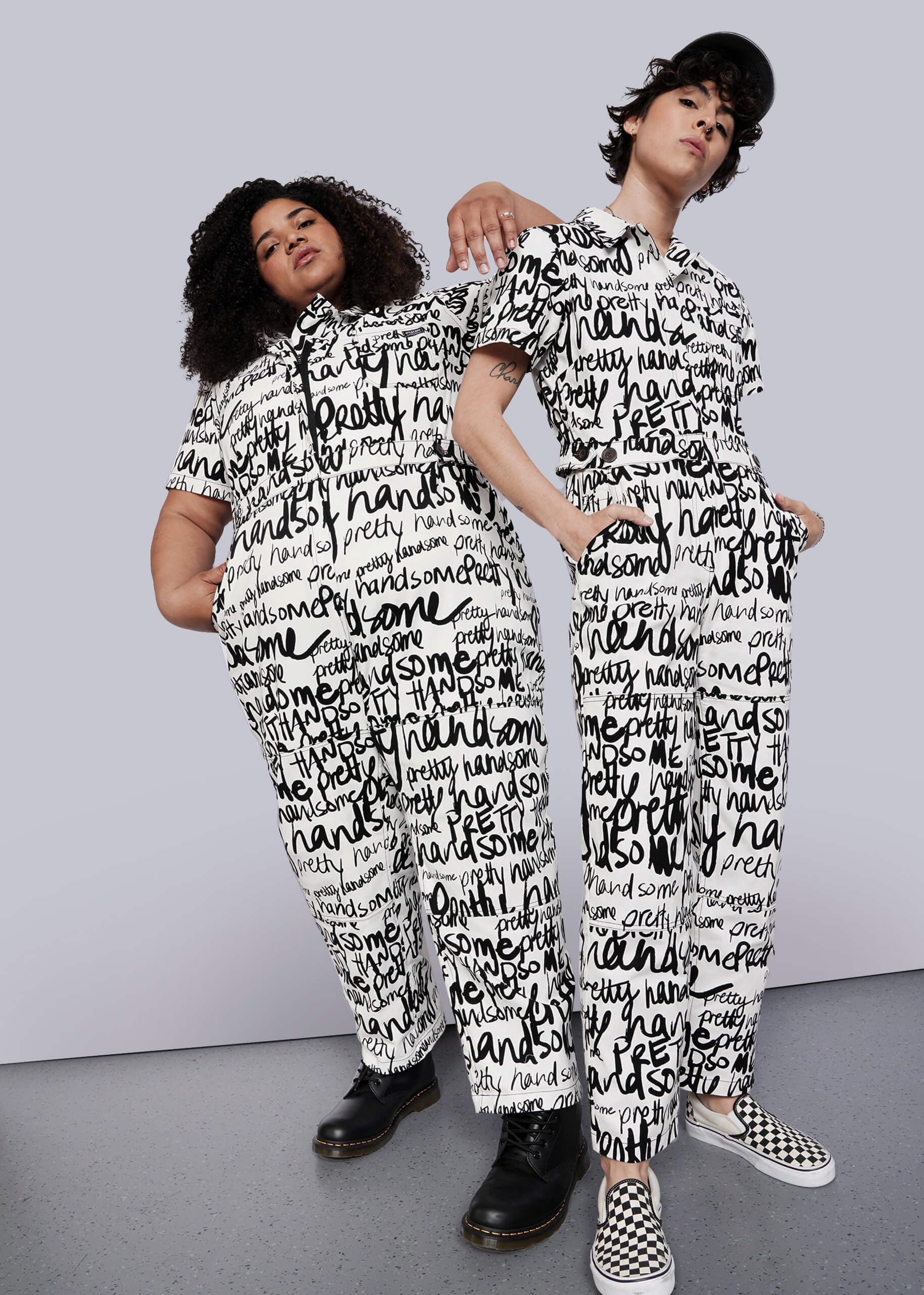 Two people pose against a plain white background in The Essential High Waisted Coverall, featuring a matching white design with bold black handwritten text. One pairs it with boots, the other with checkered slip-ons and a cap.