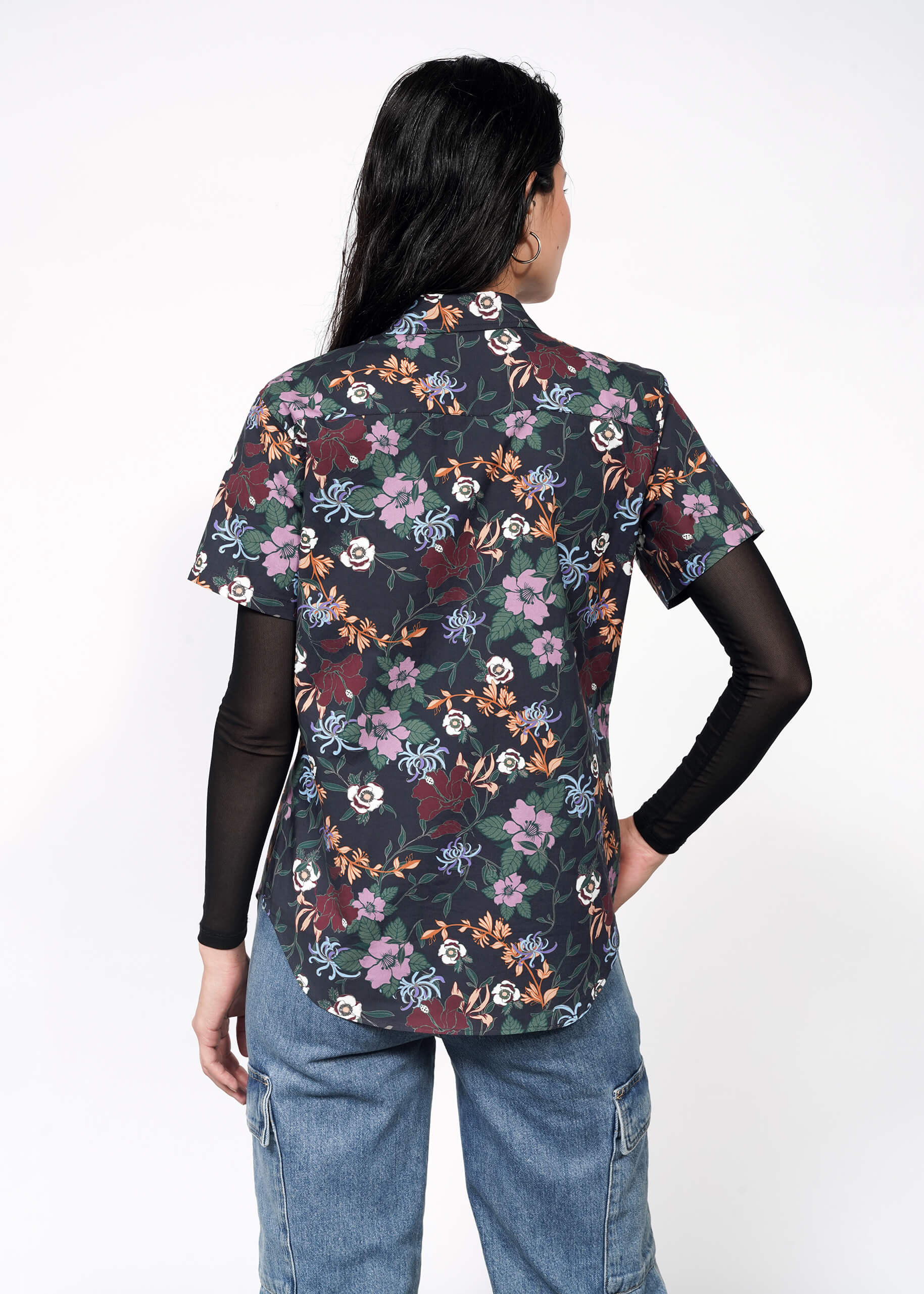 A person with long dark hair, seen from behind, wears The Essential Button Up over a black long-sleeve top and blue jeans against a plain white background.