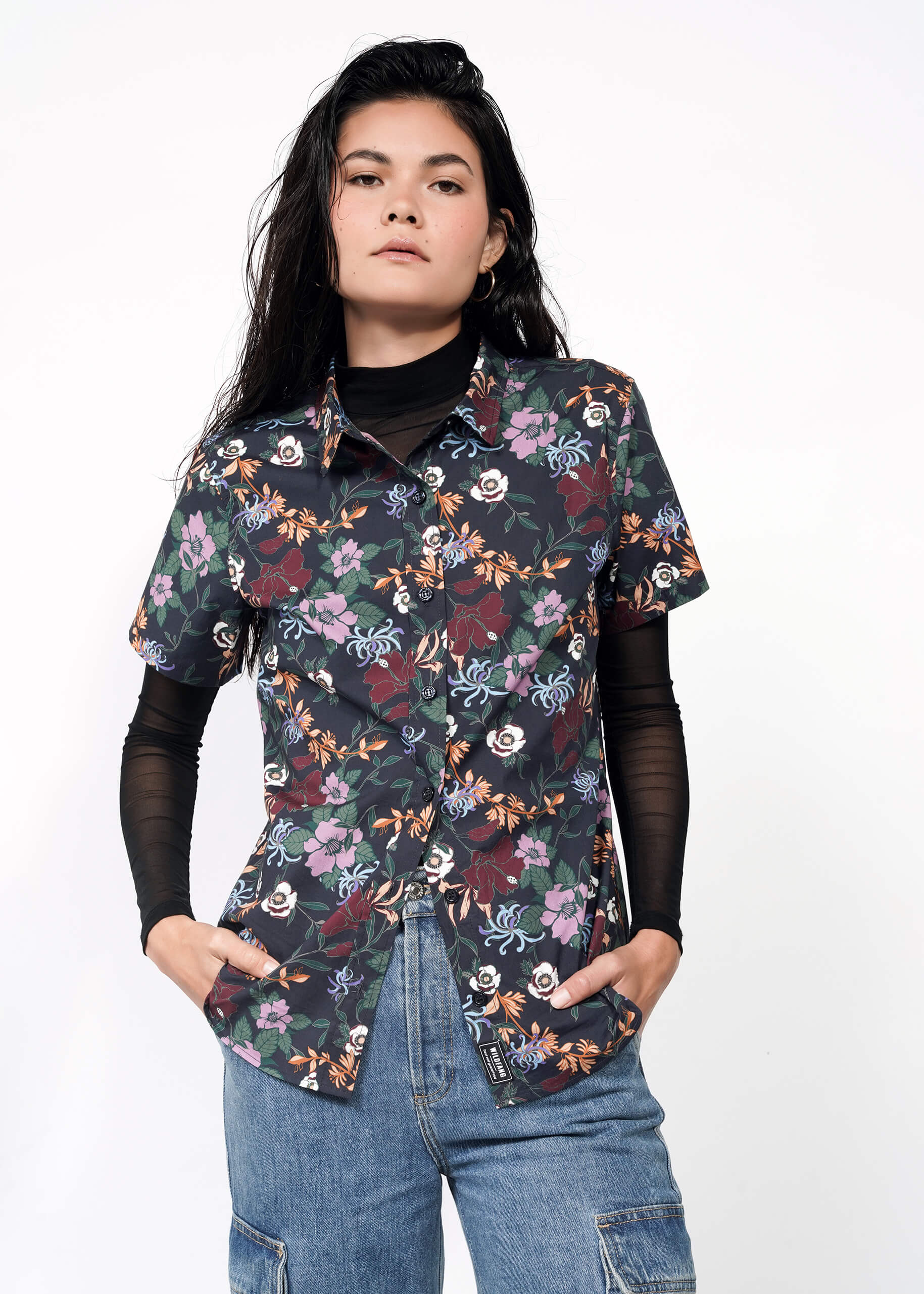 A person with long dark hair wears The Essential Button Up in black print, styled over a black long-sleeve top and blue jeans, standing with hands in pockets against a plain white background.