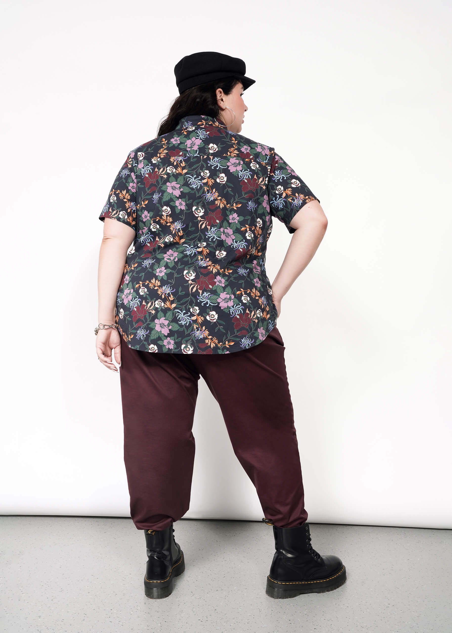 A person faces away, wearing a black cap, The Essential Button Up in dark Wildfang floral print with a tailored fit and no chest gap, burgundy pants, and black boots, one hand on their hip against a plain white background.