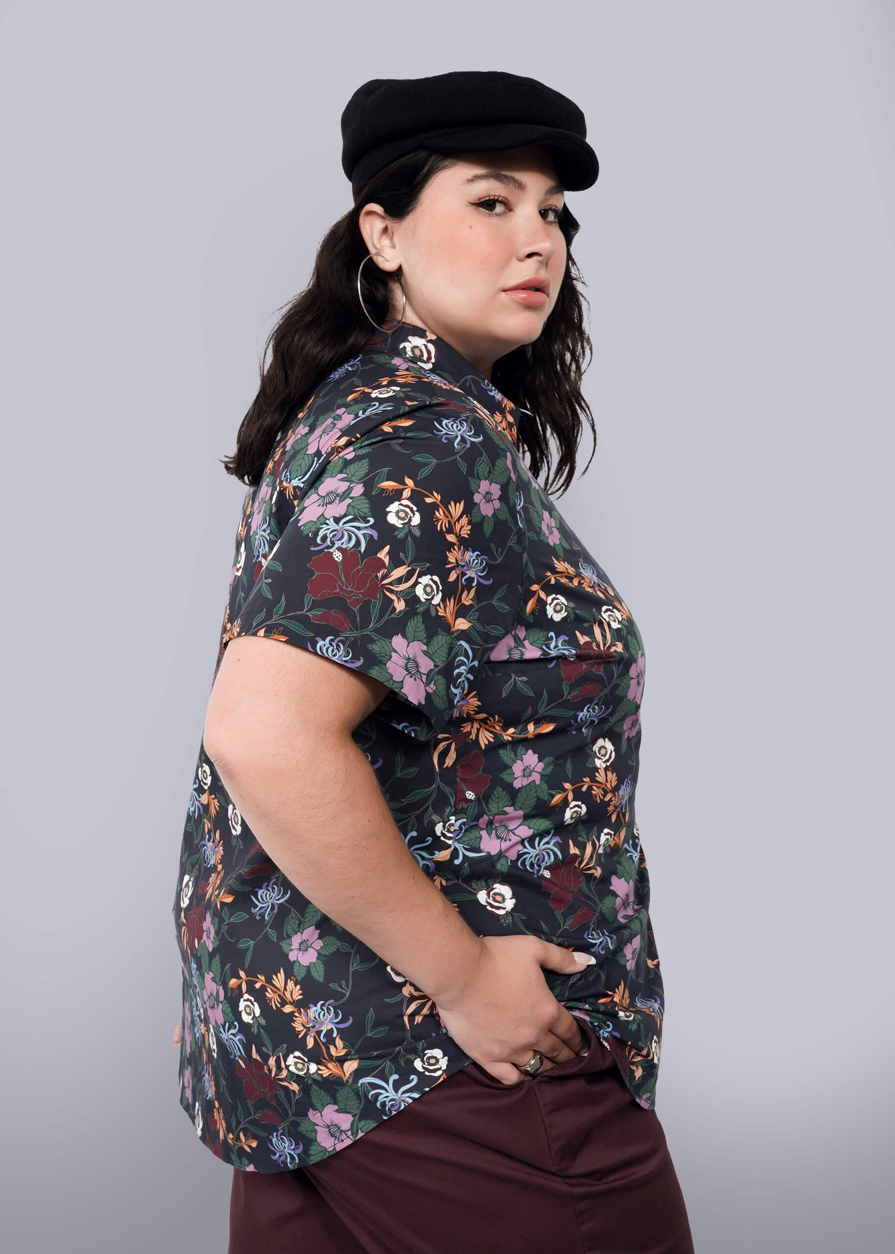 1X=A person with long dark hair wears a black newsboy cap, The Essential Button Up in a print floral short-sleeve, tailored fit with no chest gap, and maroon pants. They stand sideways to the camera against a plain white background.