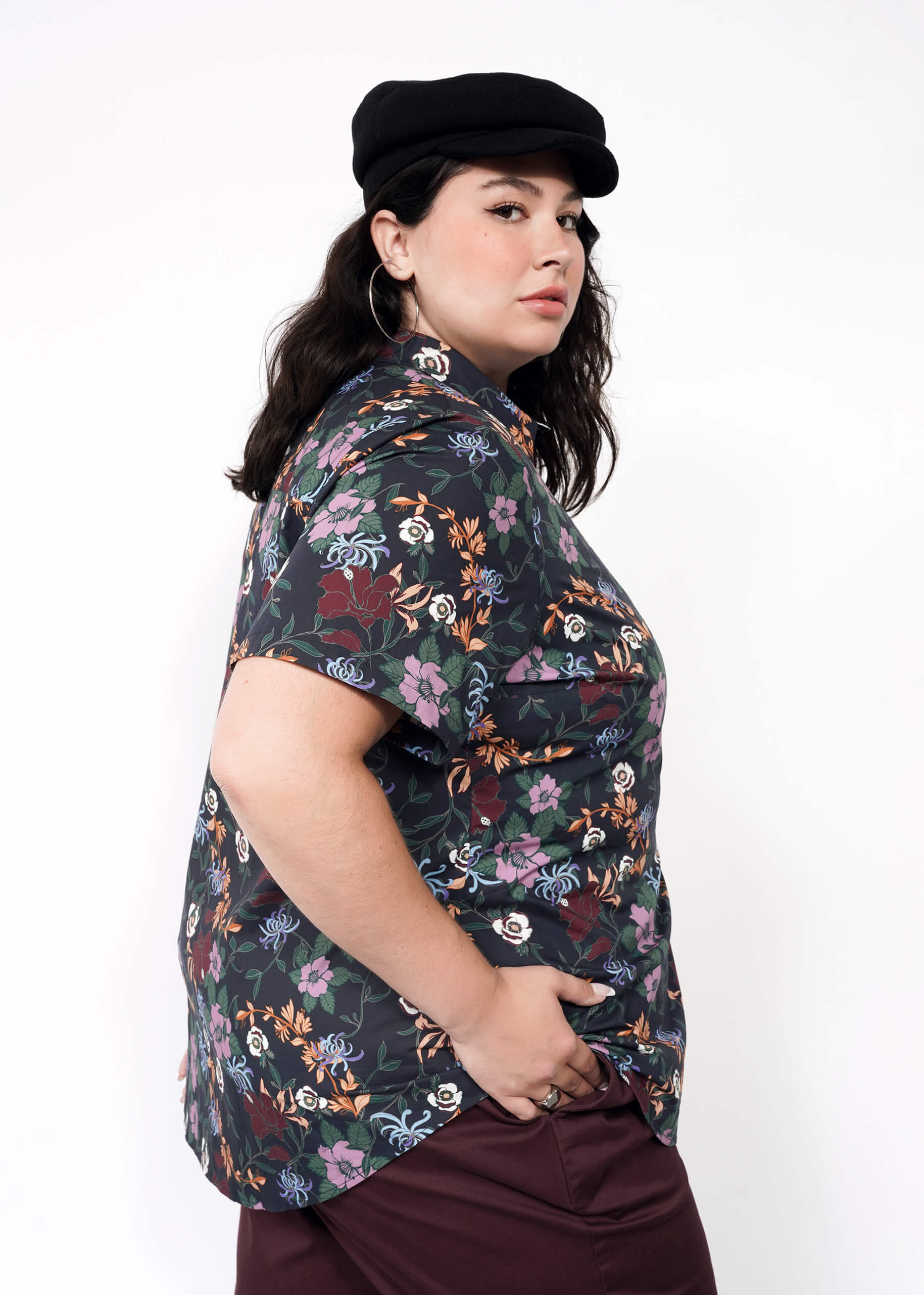 A person with long dark hair wears a black newsboy cap, The Essential Button Up in a print floral short-sleeve, tailored fit with no chest gap, and maroon pants. They stand sideways to the camera against a plain white background.