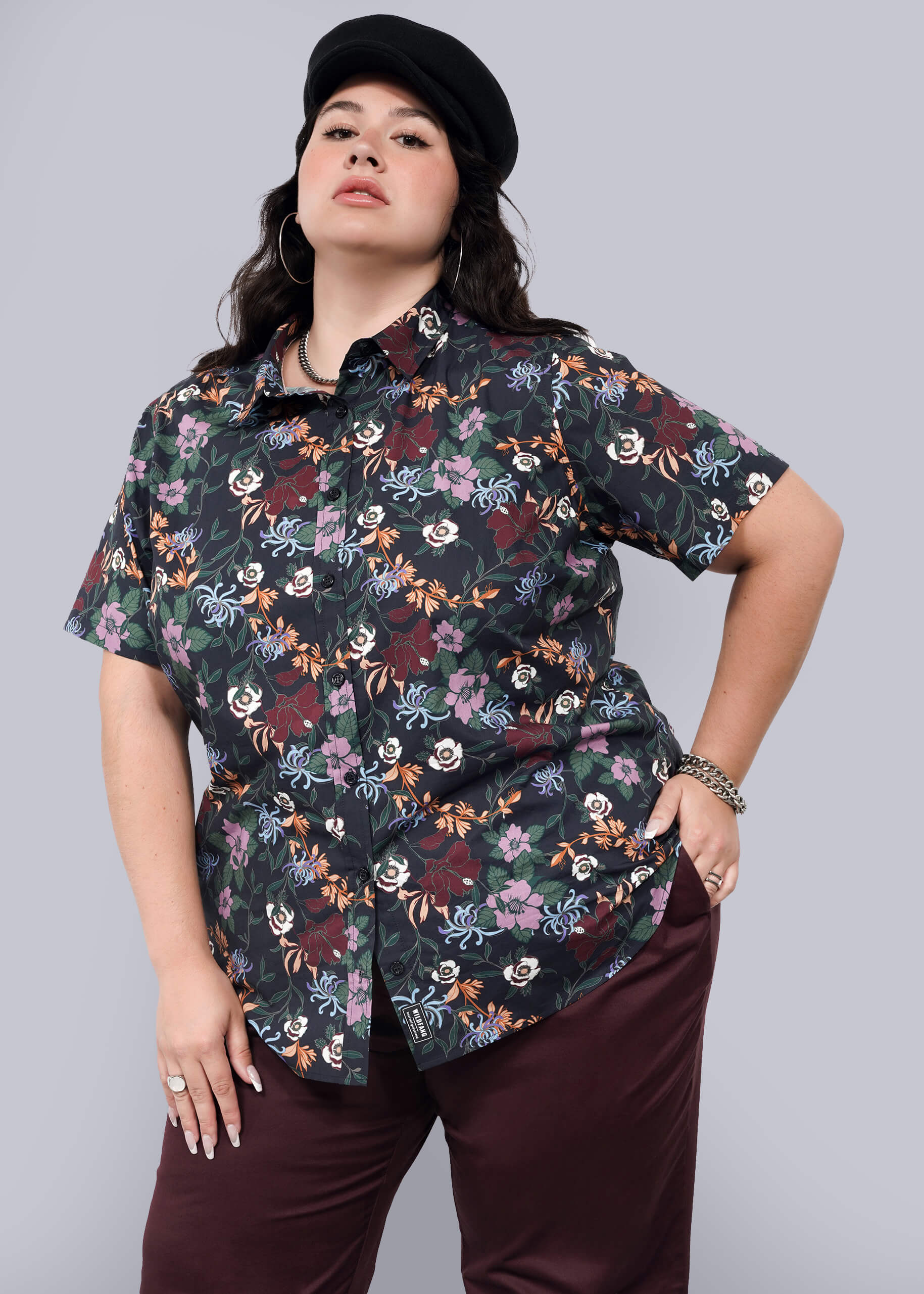 1X= A woman poses confidently against a plain light background wearing The Essential Button Up—a black floral short-sleeve, tailored-fit shirt with no chest gap—paired with dark burgundy pants, a black cap, and silver jewelry.