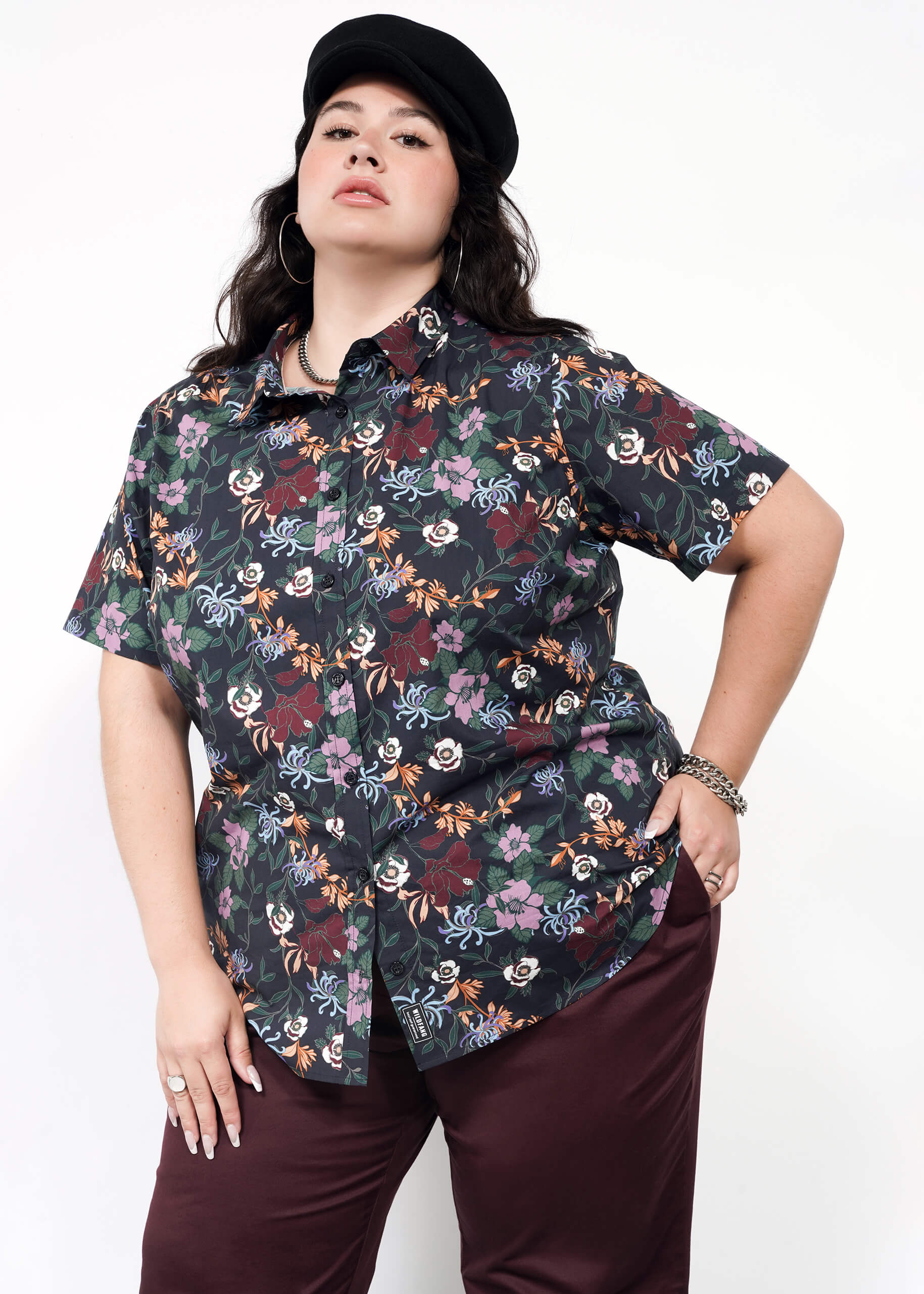 A woman poses confidently against a plain light background wearing The Essential Button Up—a black floral short-sleeve, tailored-fit shirt with no chest gap—paired with dark burgundy pants, a black cap, and silver jewelry.