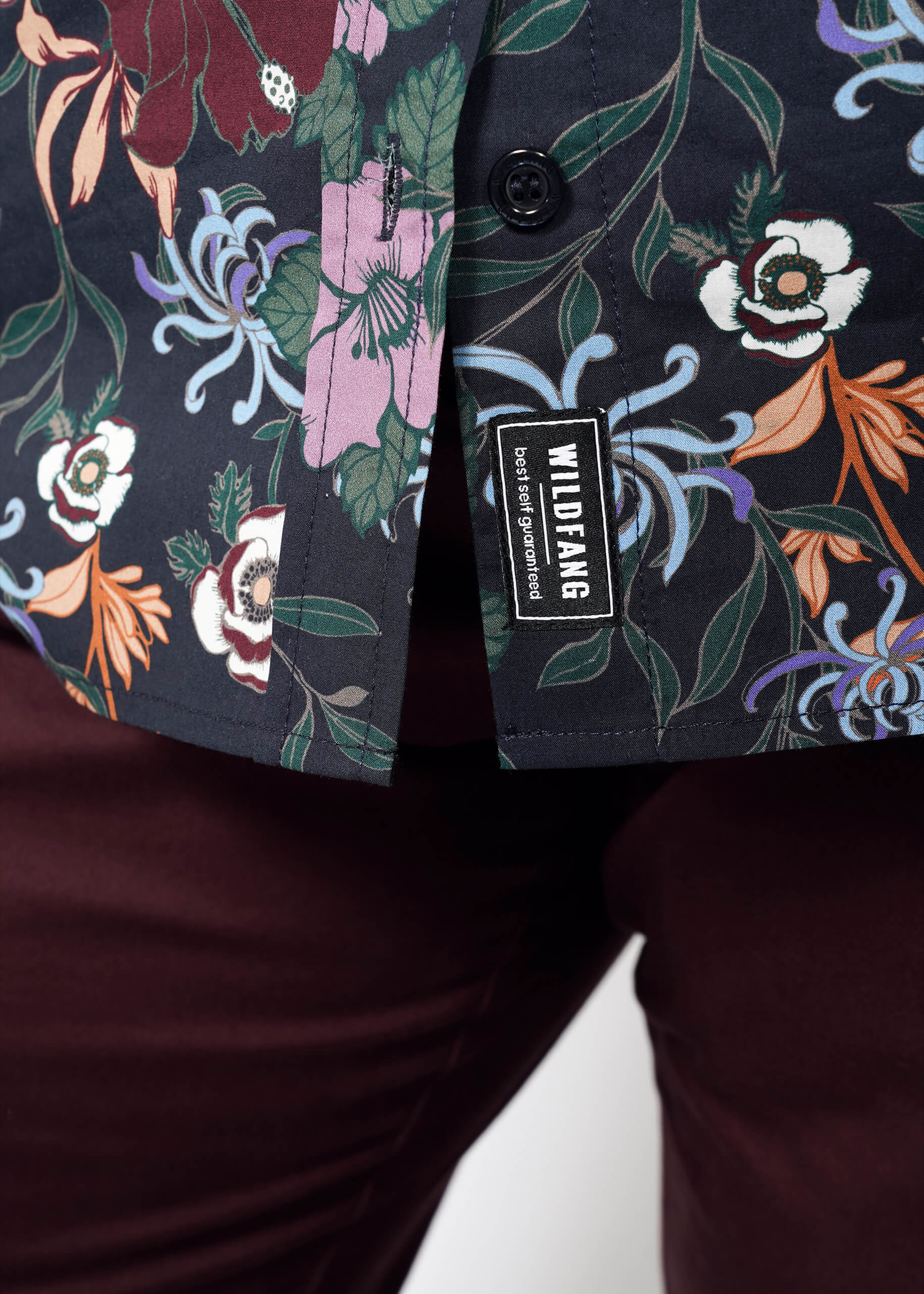 Close-up of a person wearing maroon pants and The Essential Button Up, featuring a vibrant Wildfang print. A WILDFANG brand tag is visible by the hem, showcasing the shirt’s tailored fit and attention to detail.