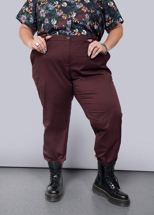 1X=A person in a floral shirt and The Essential Trouser with a burgundy, tapered leg stands with hands in pockets. They wear black boots on a grey speckled floor against a plain white background.